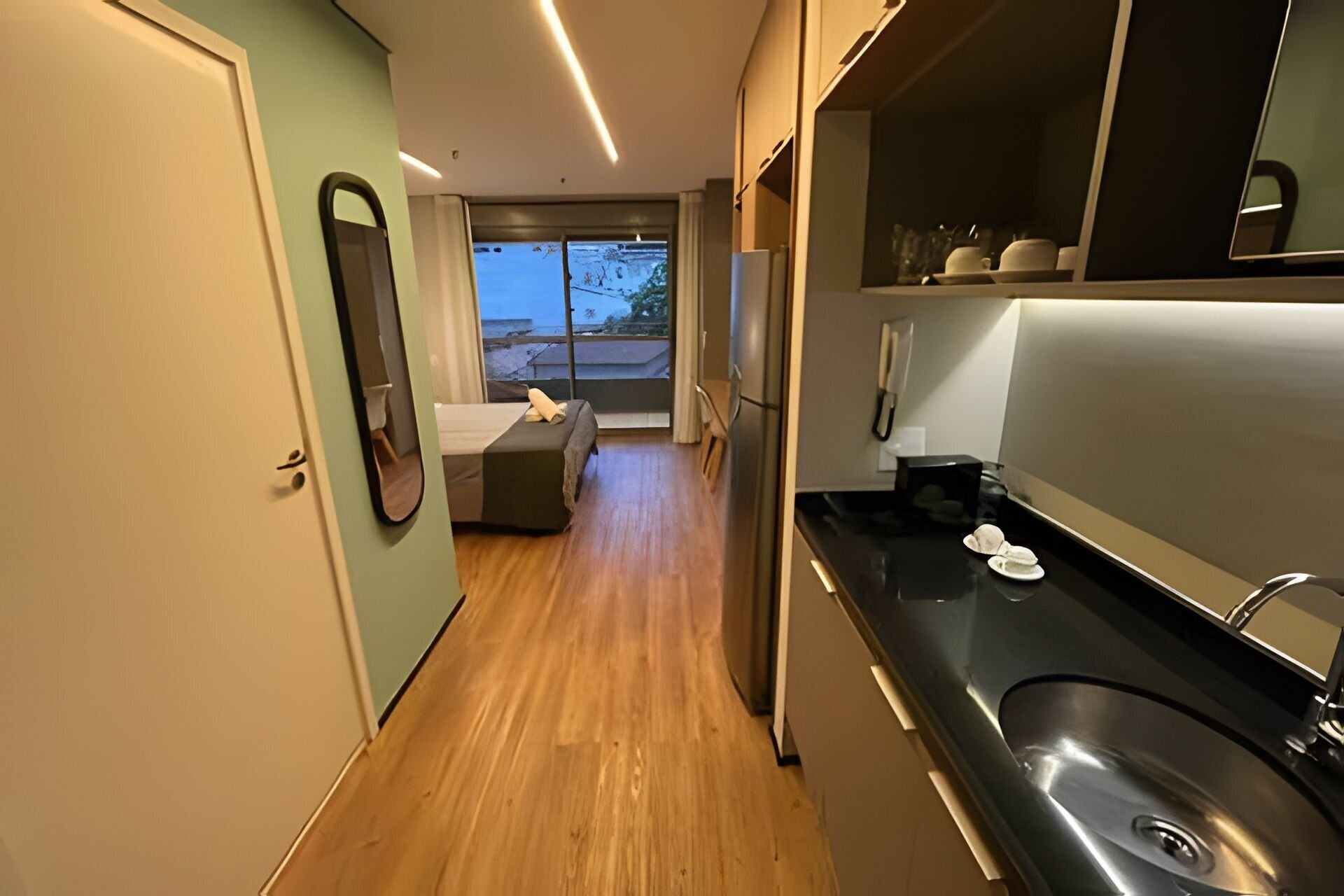 Una moderna camera d'albergo con pavimento in legno, un letto, una piccola zona salotto, una cucina con lavello e un'ampia finestra.