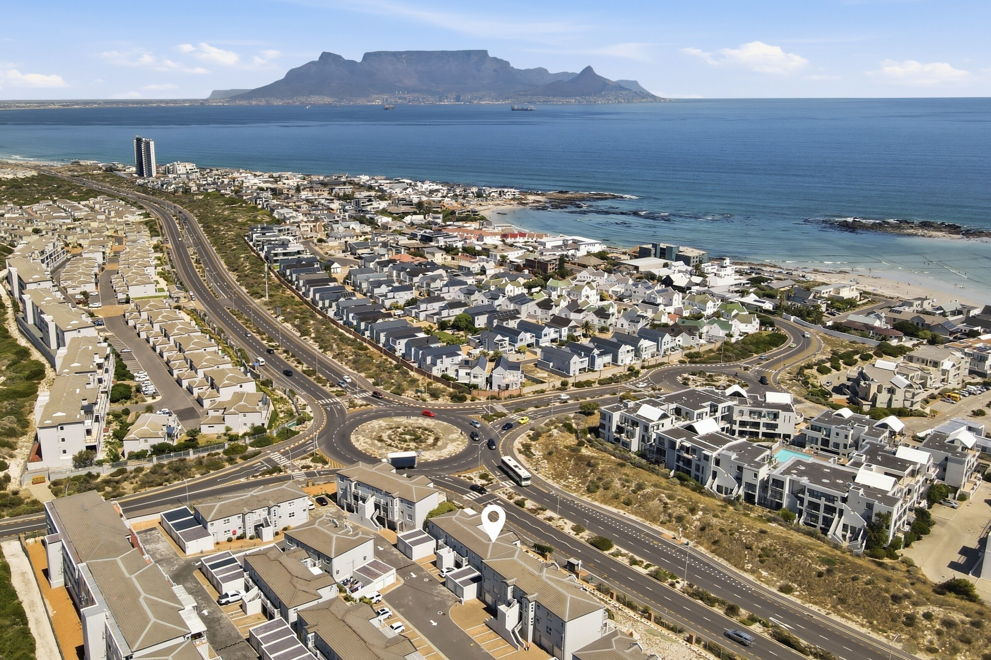An aerial view of a coastal residential area with modern housing, roads, and a prominent mountain in the background.