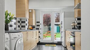 Ett kök med tvättmaskin, träskåp, kakel ovanför arbetsbänken och en utsikt genom en dörr ut mot en trädgård på baksidan.
