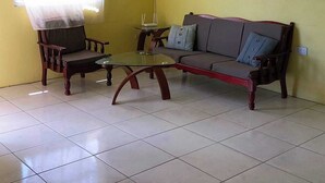 Een kamer met een gele muur, een bank, twee fauteuils, een klein tafeltje en een raam met een transparant gordijn.