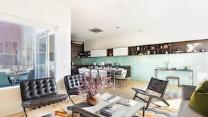 Una sala de estar moderna con un sofá, una mesa de centro y sillas. Se ve una cocina al fondo con armarios y un comedor.