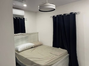 Un lit simple avec une tête de lit, une table de chevet, un luminaire suspendu et une fenêtre avec des rideaux.
