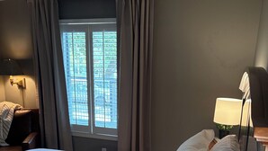 Een hotelkamer met een groot bed, een raam met gordijnen, een nachtkastje met een lamp en een wandlamp.