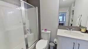 Et moderne badeværelse med brusekabine i glas, toilet, vask med spejl og en skraldespand.