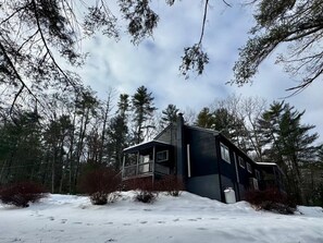 Eine zweistöckige Holzhütte mit Balkon, umgeben von Wald und verschneitem Gelände.