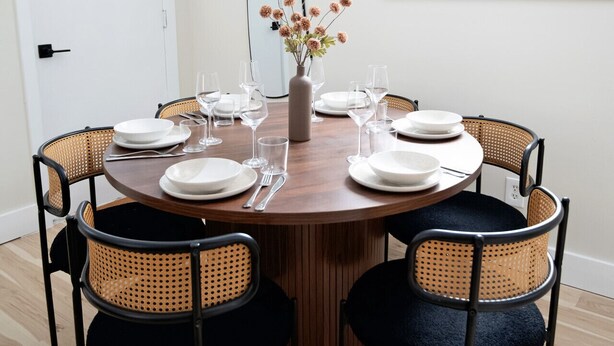A dining area with a round wooden table, six wicker chairs, and a vase with flowers. - Modern Cabin with Fire Pit | Big Bear Ski & Hiking (Eldred)