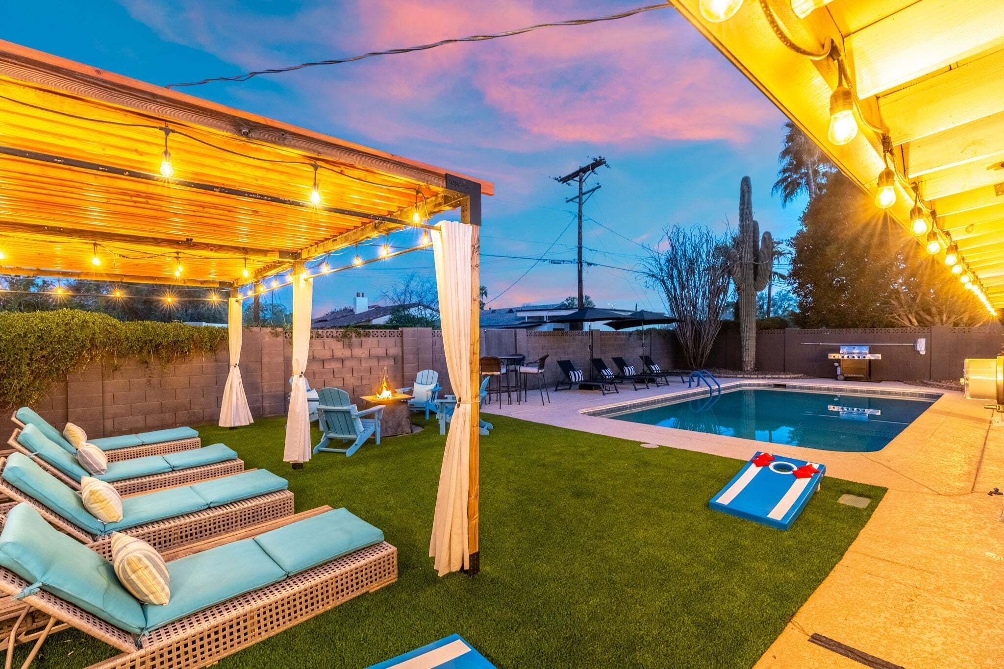 Un jardin avec une piscine, des chaises longues, une pergola illuminée de guirlandes lumineuses et un foyer extérieur.