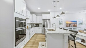 Une cuisine moderne avec des meubles blancs, des appareils en acier inoxydable, un grand îlot central doté d’un évier, des luminaires suspendus et un plancher en bois.