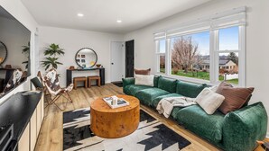 A modern living room with a green sofa, a wooden coffee table, and a large window with a view of houses.