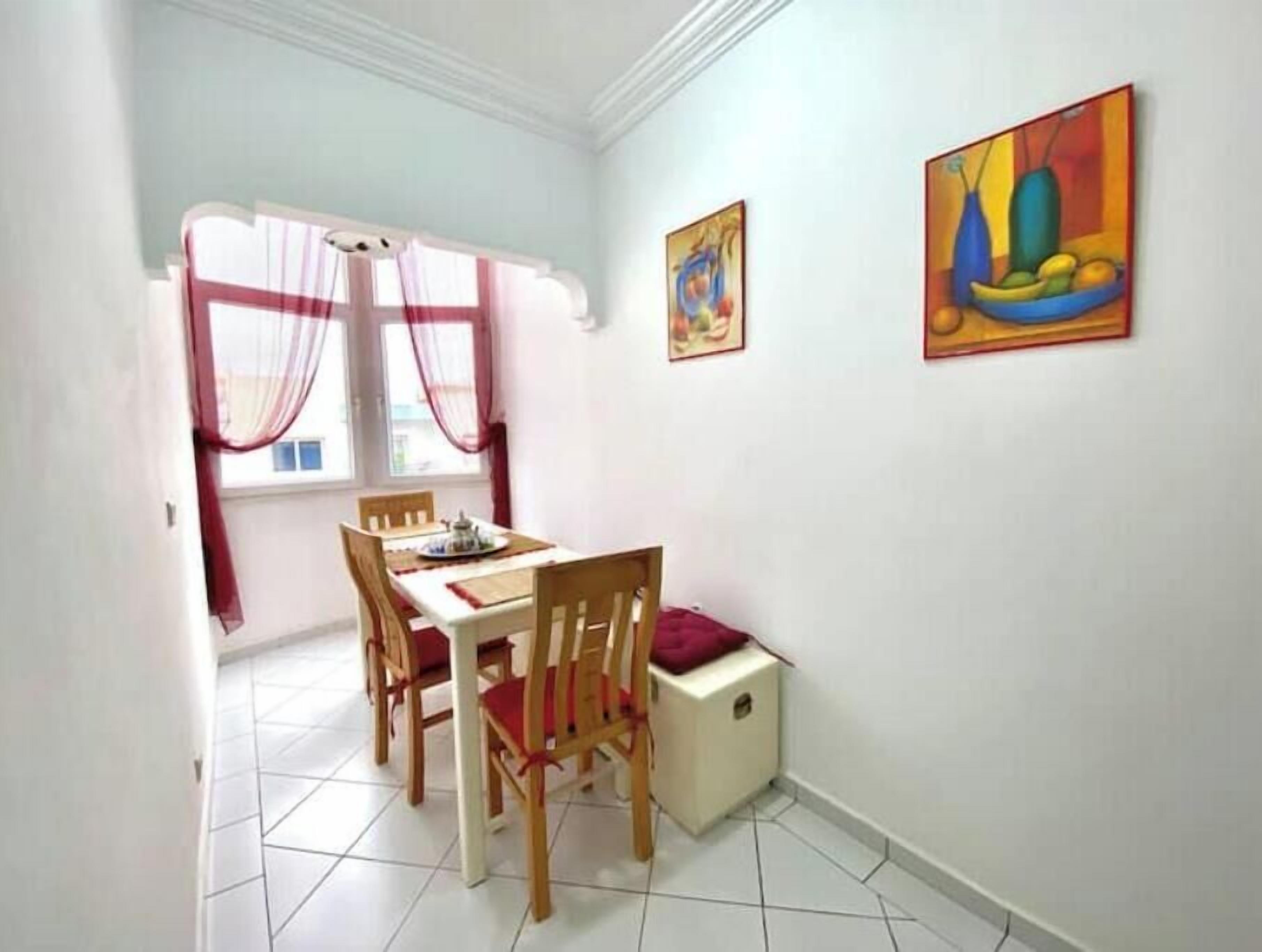 A dining area with a table and chairs, a window with curtains, and two paintings on the wall.