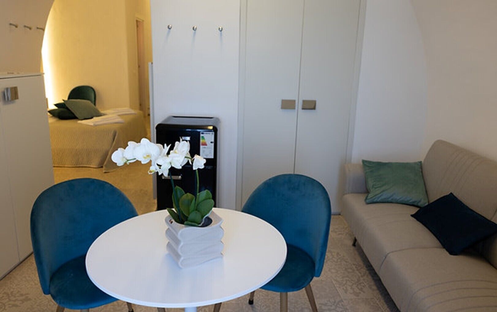 Ein Zimmer mit einem runden weißen Tisch, zwei blauen Stühlen, einem beigen Sofa und einem kleinen weißen Kühlschrank.