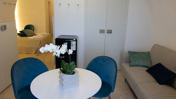 Ein Zimmer mit einem runden weißen Tisch, zwei blauen Stühlen, einem beigen Sofa und einem kleinen weißen Kühlschrank.