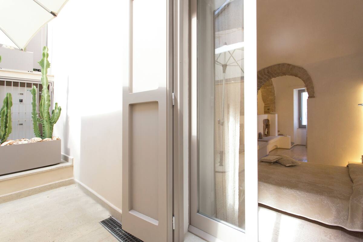 Ein modernes Interieur mit einer Schiebetür aus Glas, einem Bett mit Kopfteil, einem Steingewölbe und einer Pflanzenecke mit Kakteen.