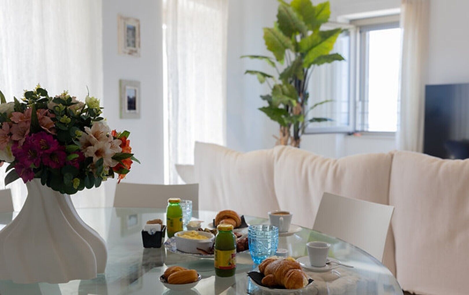 A dining table with food, drinks, and a vase of flowers. There is a sofa and a plant in the background.
