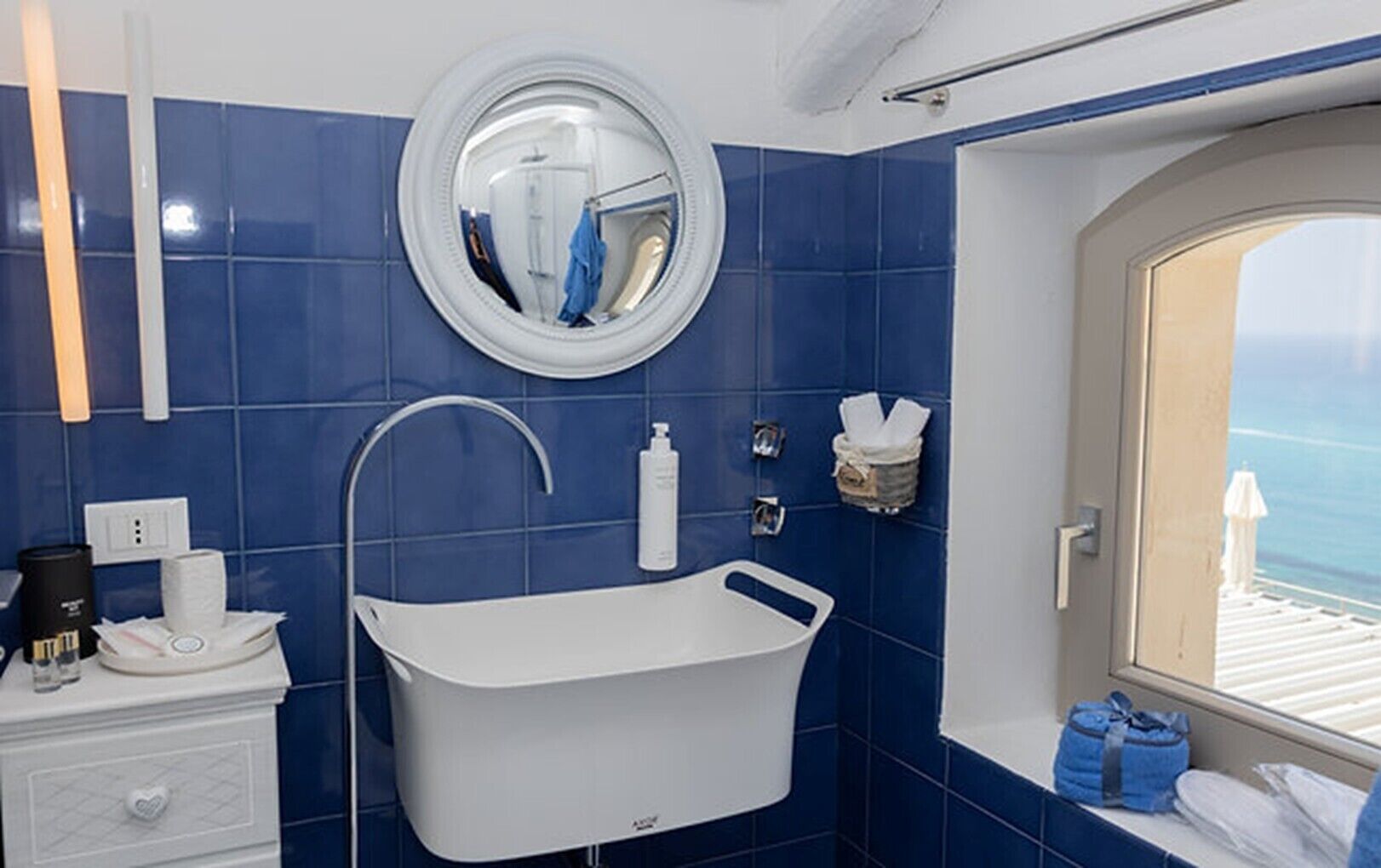 A bathroom with a bathtub, a round mirror, a towel rack, and a window with a view.