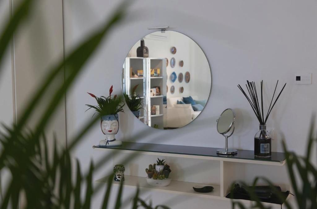 A mirror on a wall with a shelf below it containing a small plant, a decorative item, and a bottle of reed diffuser.
