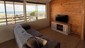 Un salon moderne avec une grande fenêtre, un canapé, un téléviseur posé sur un meuble en bois et une vue sur la mer.