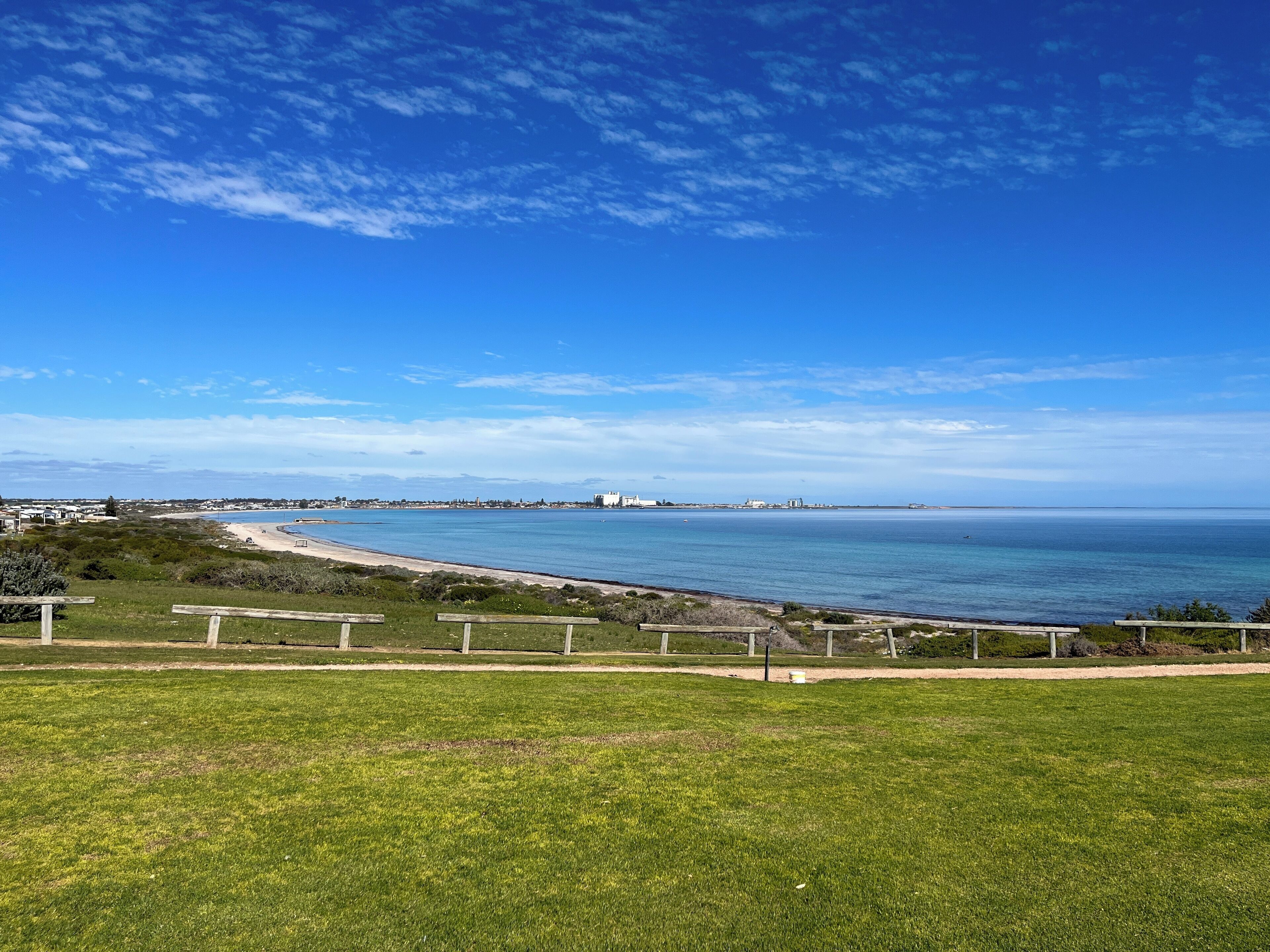 Eine Küstenlandschaft mit einer grünen Fläche, einem Strand und Blick auf das Meer sowie entfernte Gebäude.
