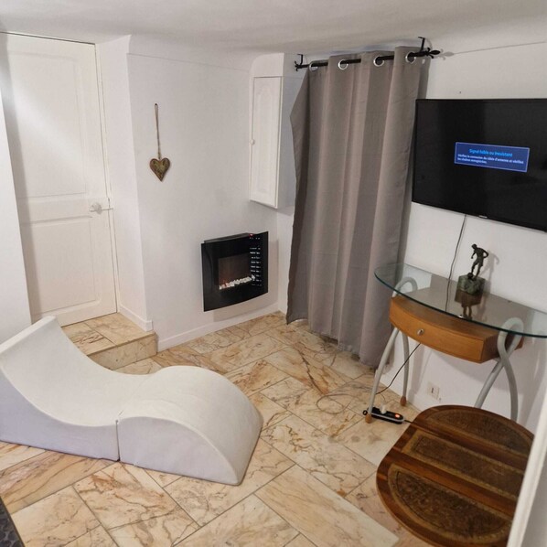 A room with a white curved bench, a small fireplace, a glass table with a wooden base, and a television mounted on the wall.