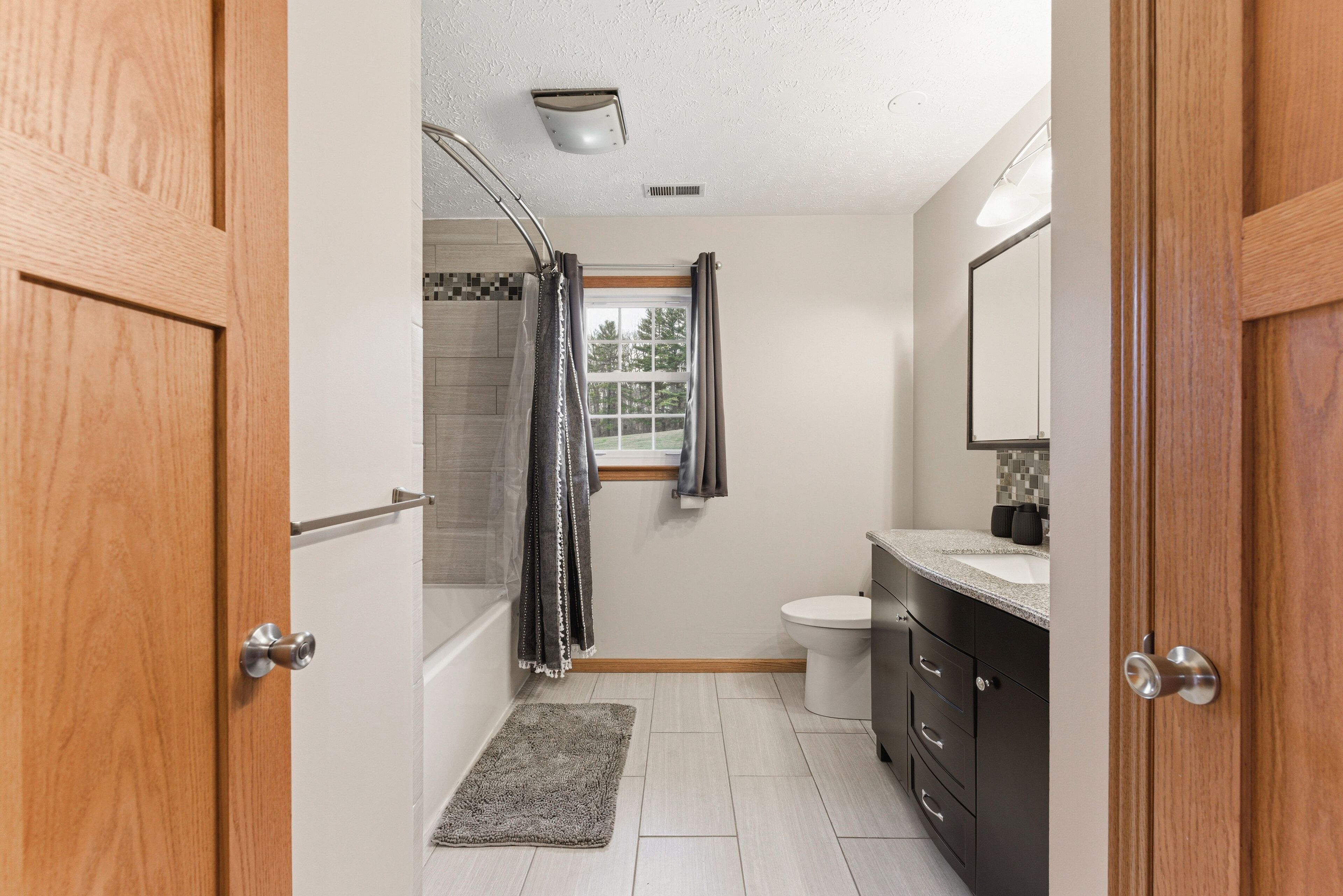 A bathroom with a shower, toilet, vanity, and a window with curtains.
