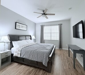 Quarto com cama, mesas de cabeceira, escrivaninha, ventoinha no teto, janela com cortinas e televisão presa na parede.