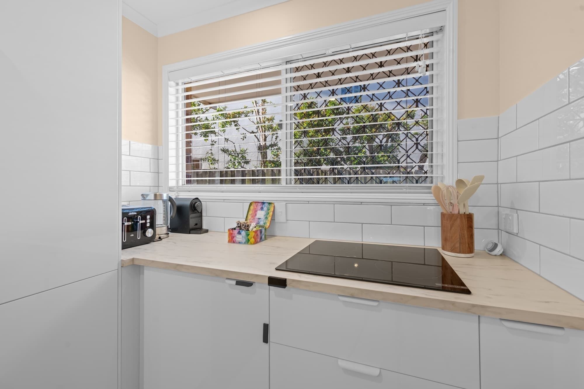 Una cocina con gabinetes blancos, encimera negra, ventana con persianas y vista a árboles.