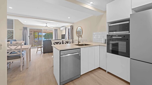 A modern kitchen with a dishwasher, oven, and a dining area with a table and chairs. - Family Friendly 2 Bed Flat with Balcony (Mooloolaba)