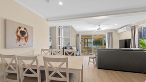 A modern living room with a sofa, dining table, and chairs. There is a large window with curtains, a ceiling fan, and an air conditioning unit. - Family Friendly 2 Bed Flat with Balcony (Mooloolaba)