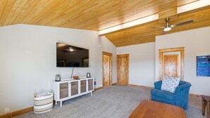 Un salón amplio con techo alto, televisor montado, sofá azul, mesa de centro de madera y un mueble blanco.