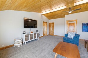 Ein geräumiges Wohnzimmer mit hoher Decke, einem Wand-Fernseher, einem blauen Sofa, einem Holztisch und einer weißen Kommode.
