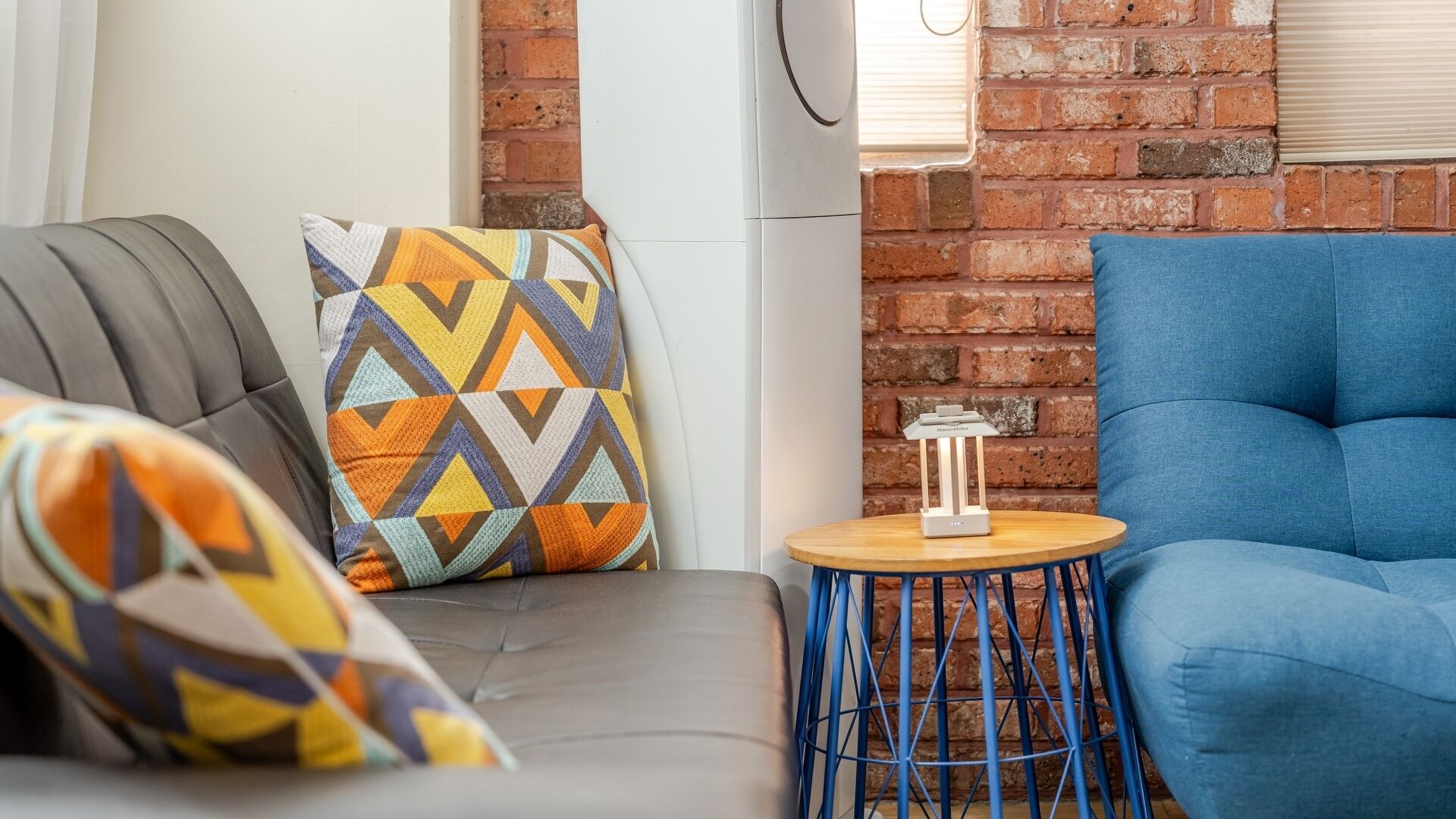 A modern living room with a grey sofa, a blue armchair, and a patterned throw pillow. A small wooden table with a white lamp is visible.