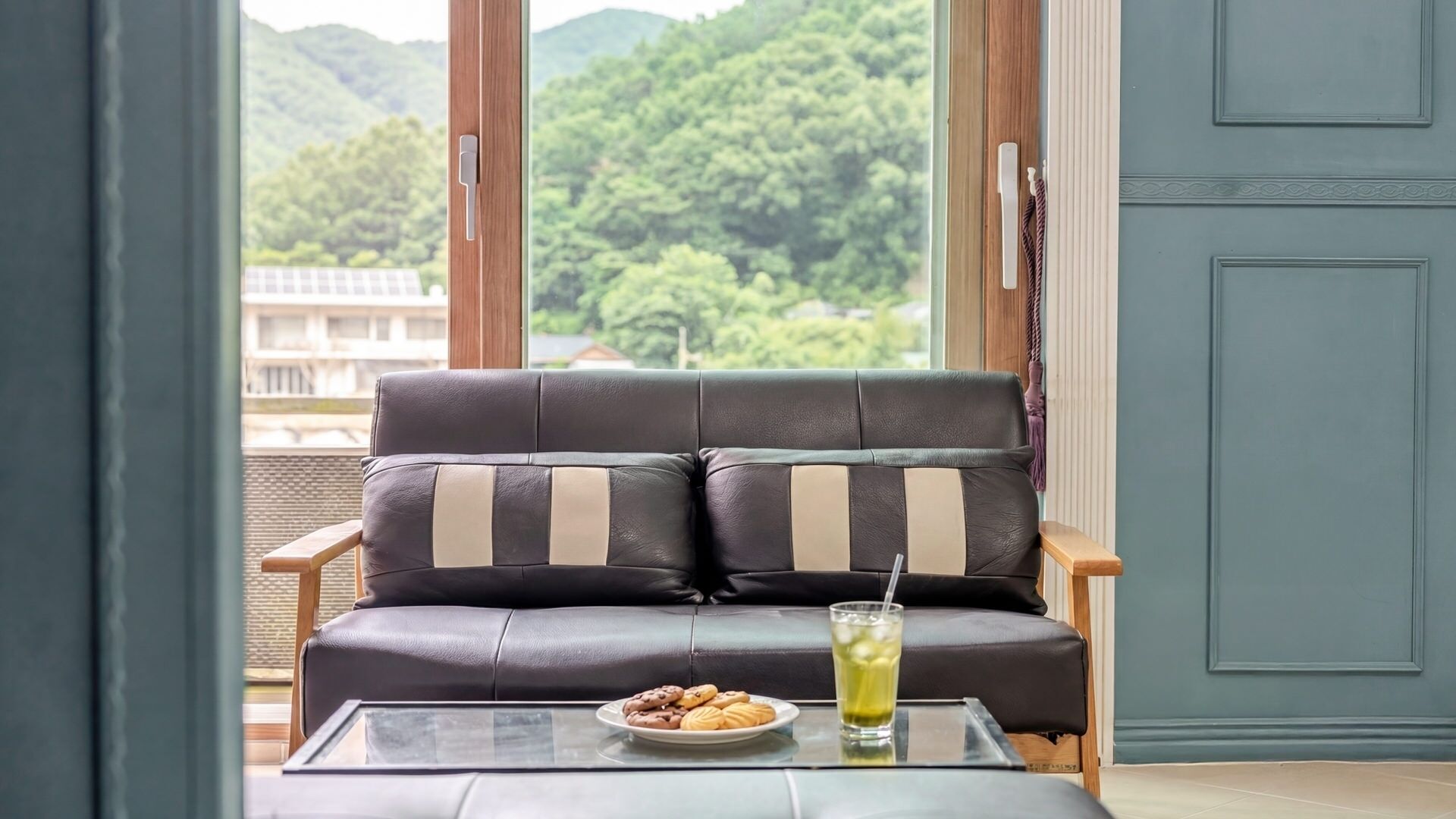 Un sofá de cuero con cojines a rayas, una mesa de vidrio con un plato de galletas y un vaso de bebida, y una vista de vegetación a través de una ventana.