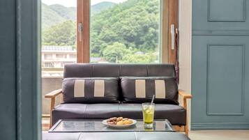 Un sofá de cuero con cojines a rayas, una mesa de vidrio con un plato de galletas y un vaso de bebida, y una vista de vegetación a través de una ventana.