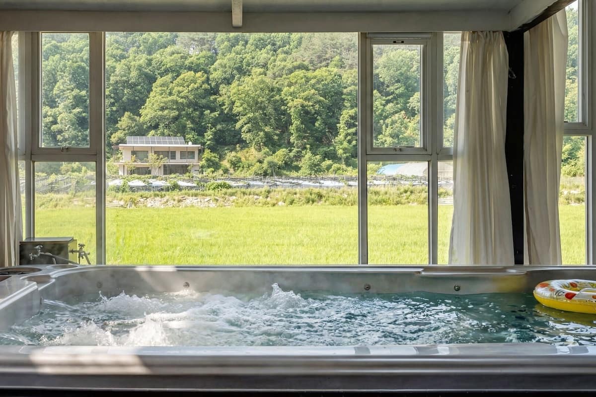 Un bain Ă remous avec vue sur un champ verdoyant et des arbres, grĂące Ă de grandes fenĂȘtres.
