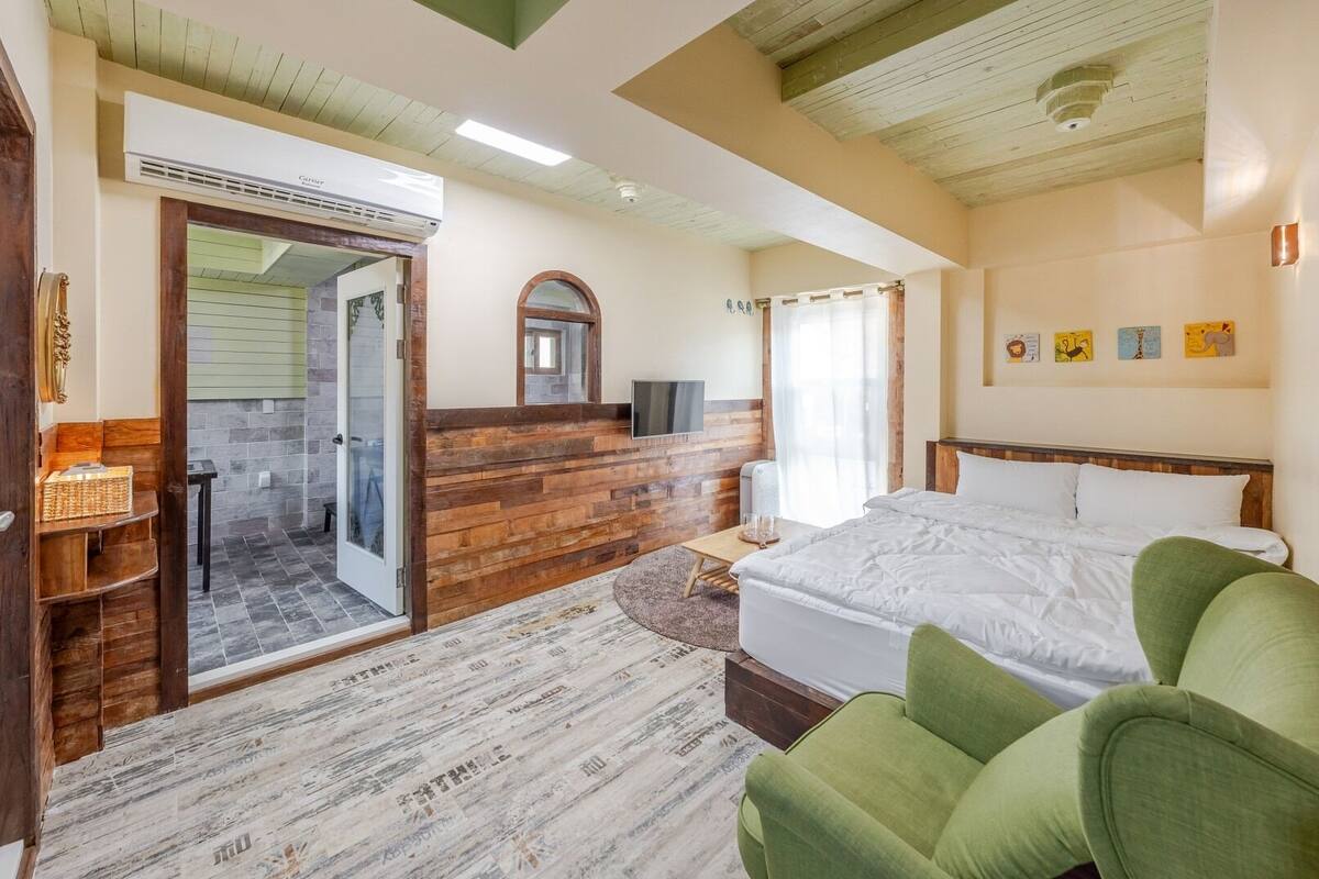 Ein Schlafzimmer mit Bett, einem grünen Sessel, einer Holzwand, einem Fernseher und einem Badezimmer mit Dusche und Toilette.
