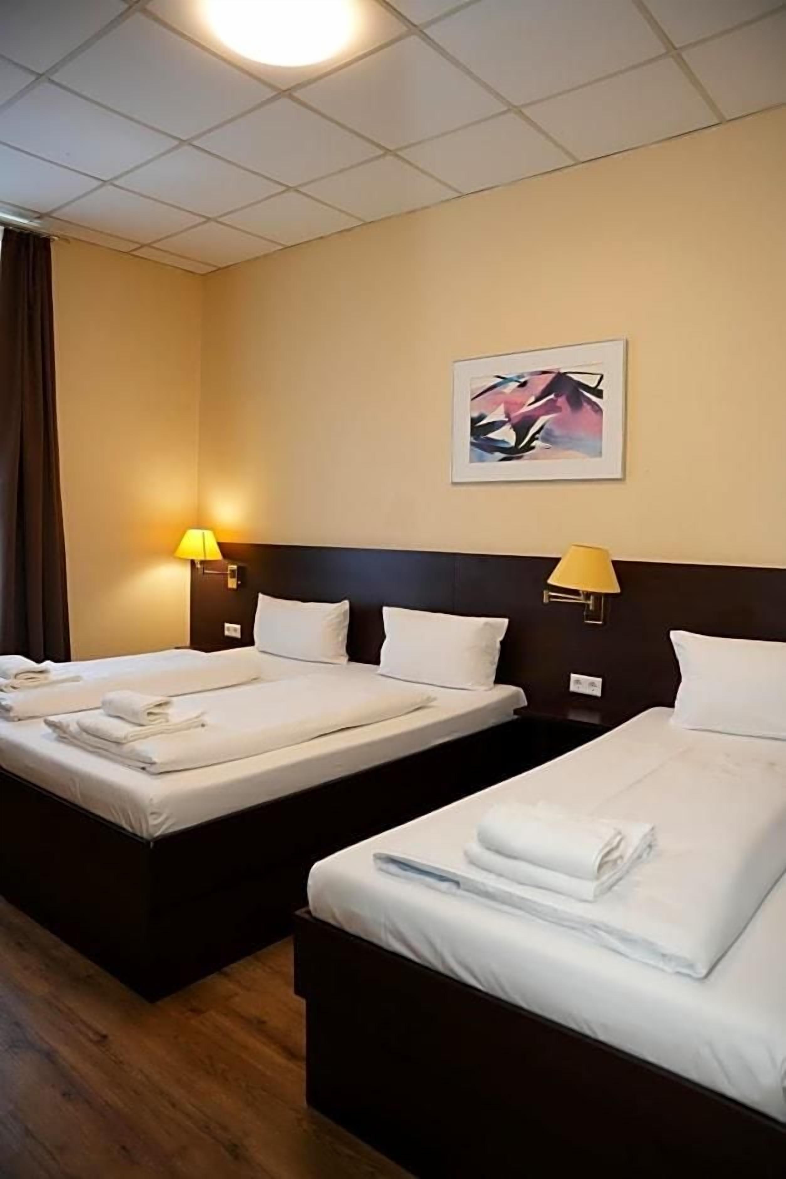 A hotel room with two beds, each with a pillow and a folded towel. There is a wall-mounted lamp on each side of the beds.