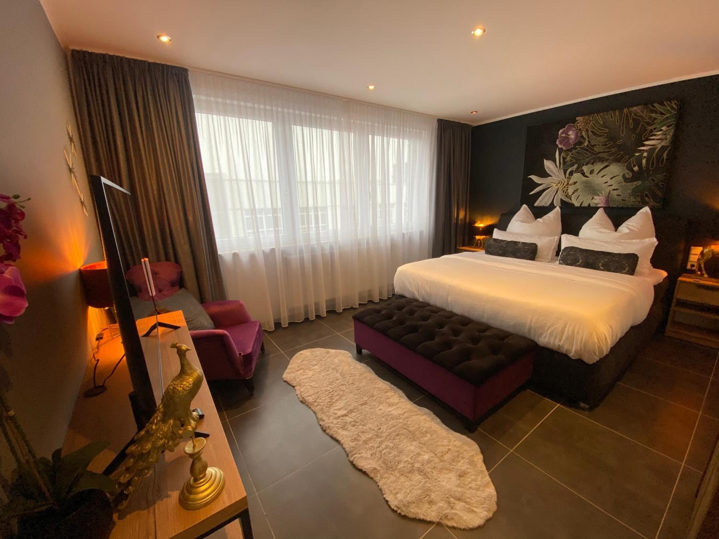 Un dormitorio moderno con una cama grande, un sillón, una mesita de noche con lámpara, y un ventanal con cortinas traslúcidas.