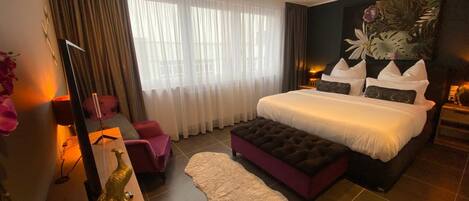 Un dormitorio moderno con una cama grande, un sillón, una mesita de noche con lámpara, y un ventanal con cortinas traslúcidas.