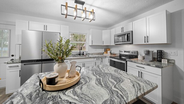 Una cocina moderna con armarios blancos, encimera de granito, electrodomésticos de acero inoxidable y una isla central con planta y reloj.