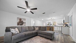 Una sala de estar moderna con un sofá modular, un ventilador de techo y una cocina al fondo.