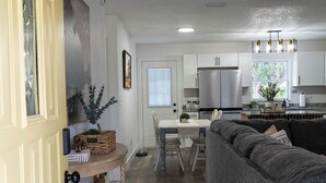 Una cocina moderna con electrodomésticos de acero inoxidable, un comedor con un sofá gris y una mesita, y un pasillo con una puerta de madera y cestas.