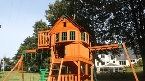 A wooden play structure with a house-like design, including a swing set and a slide. - Latitude Ouest Hotel Restaurant Spa (Locronan)