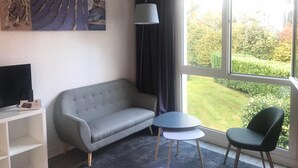 A room with a grey sofa, a small round table, two chairs, a TV on a stand, and a window with a view of a garden. - Latitude Ouest Hotel Restaurant Spa (Locronan)