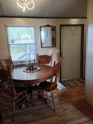 A dining room with a round wooden table, six chairs, a chandelier, a window with blinds, a mirror, and a door with a chain. - Lovely Home (mobile) bring the family to the Equestrian events in Aiken, SC! (Aiken)
