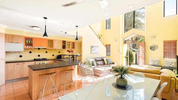 Eine moderne Küche mit Kochinsel, ein Essbereich mit Glastisch und ein Wohnzimmer mit Sofa und Pflanze.