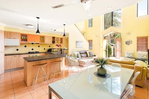 A modern kitchen with a central island, a dining area with a glass table, and a living room with a sofa and a plant.
