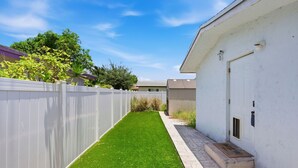 A narrow, well-maintained grassy area with a white fence, a paved walkway, and a building with a door and window. - Walk to Downtown Delray and beach.   Huge fenced-in yard for dog (Delray Beach)