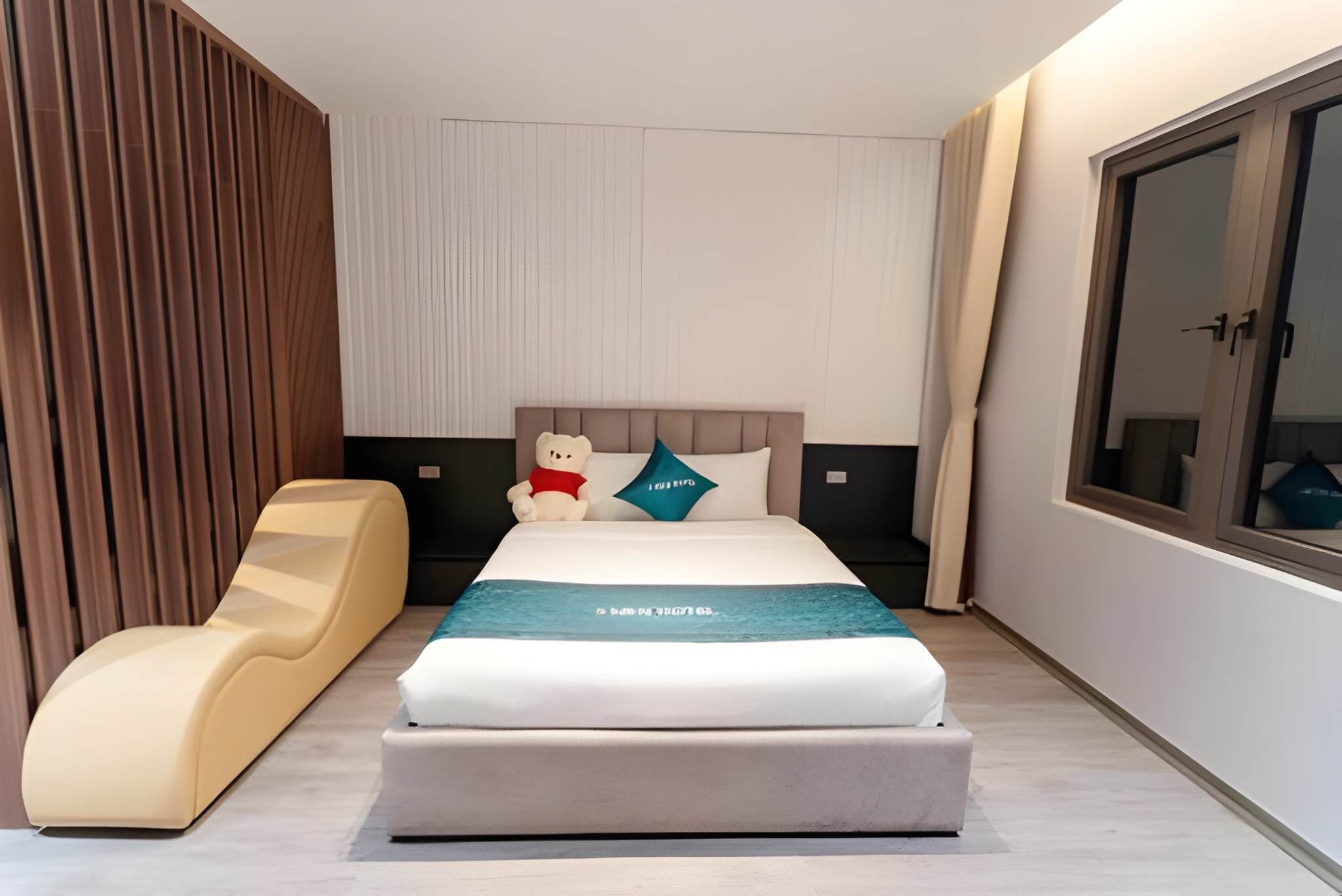 A modern hotel room with a bed, a lounge chair, a large window, and a wardrobe.