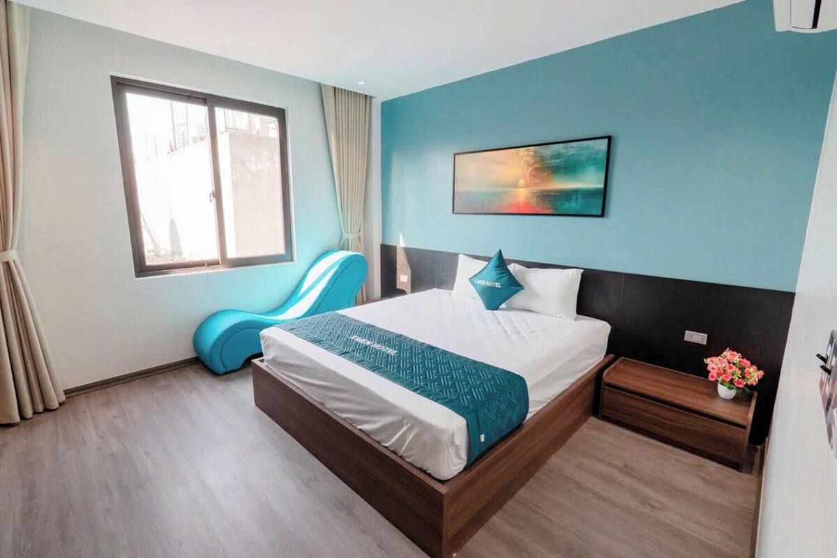 Ein Hotelzimmer mit Bett, Fenster, Stuhl, Nachttisch und einem Gemälde an der Wand.