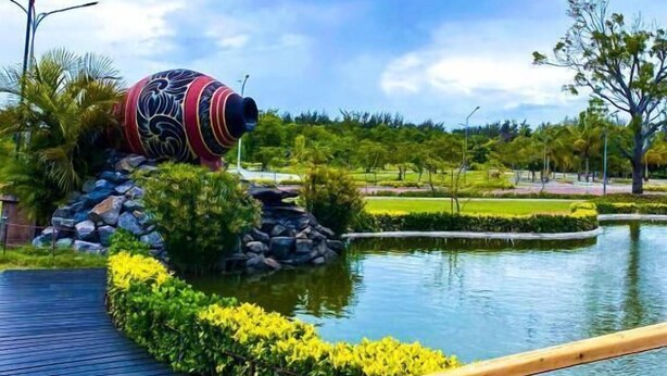 Un sentiero conduce a uno stagno con una grande scultura decorativa, circondato da vegetazione e un cielo sereno.
